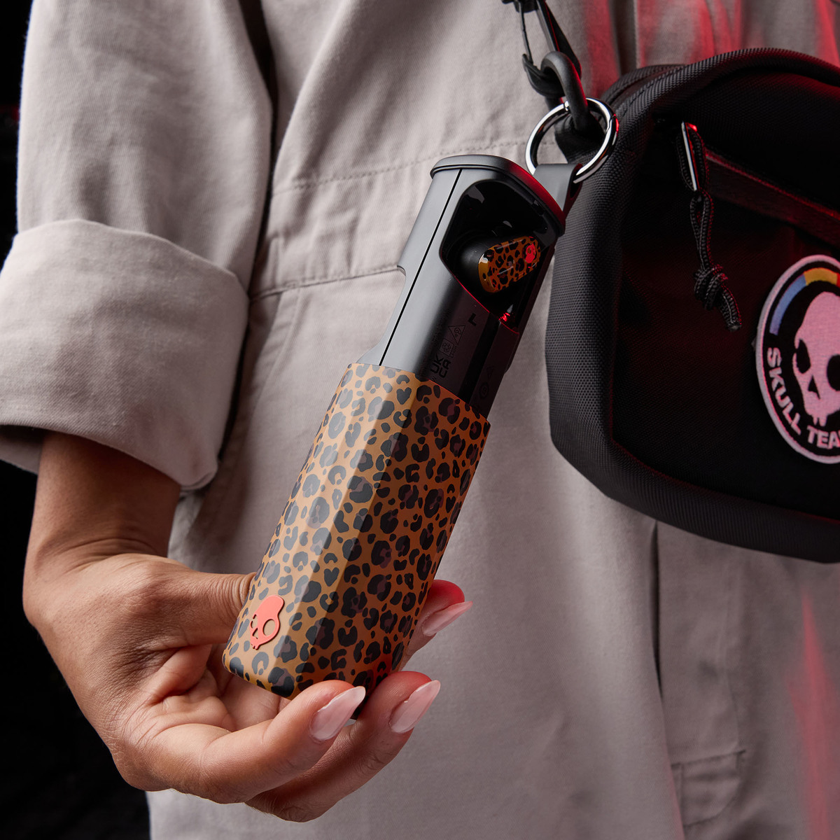 Eine Person hält ein Kopfhörer-Etui mit Leopardenmuster. Eine schwarze Tasche mit Schädel-Logo hängt an der Schulter. Rote Beleuchtung.