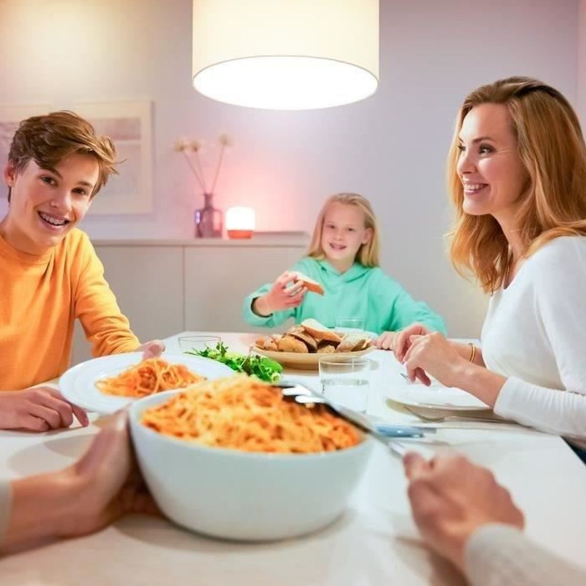 Familia cenando. Espaguetis en un bol, platos de pasta, pan y vasos de agua. Rostros sonrientes. Una lámpara cuelga sobre la mesa.