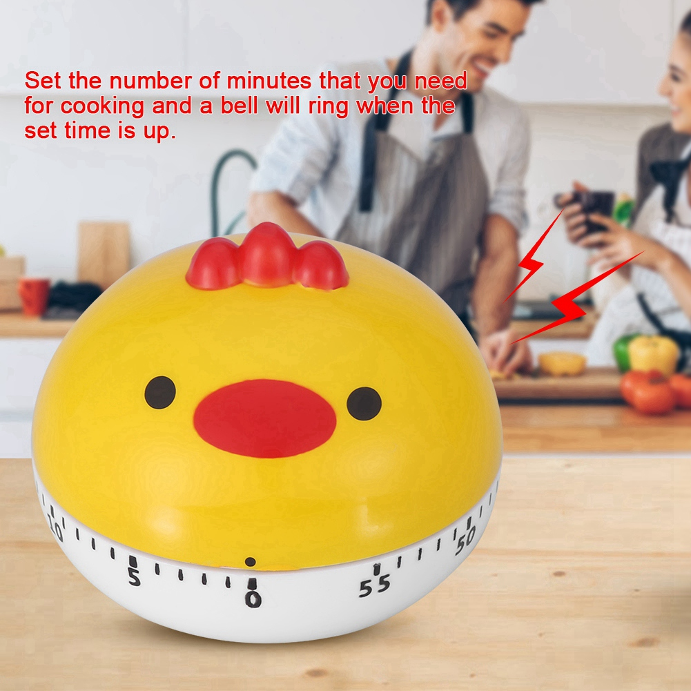 Ein gelber, hühnerförmiger Küchentimer steht auf einem Holztisch. Im Hintergrund sind zwei Personen, eine hält eine Tasse.
