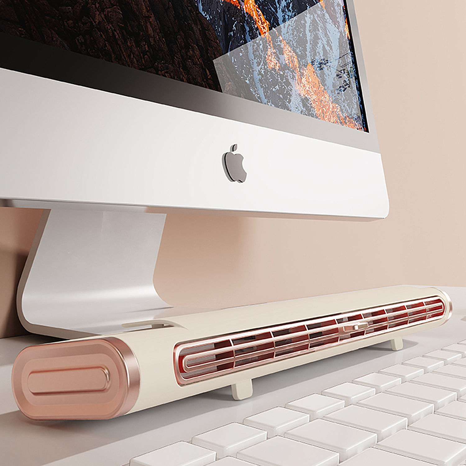Un monitor per computer con logo Apple, una tastiera bianca e un termoventilatore beige e color rame. Lo sfondo è rosa chiaro.
