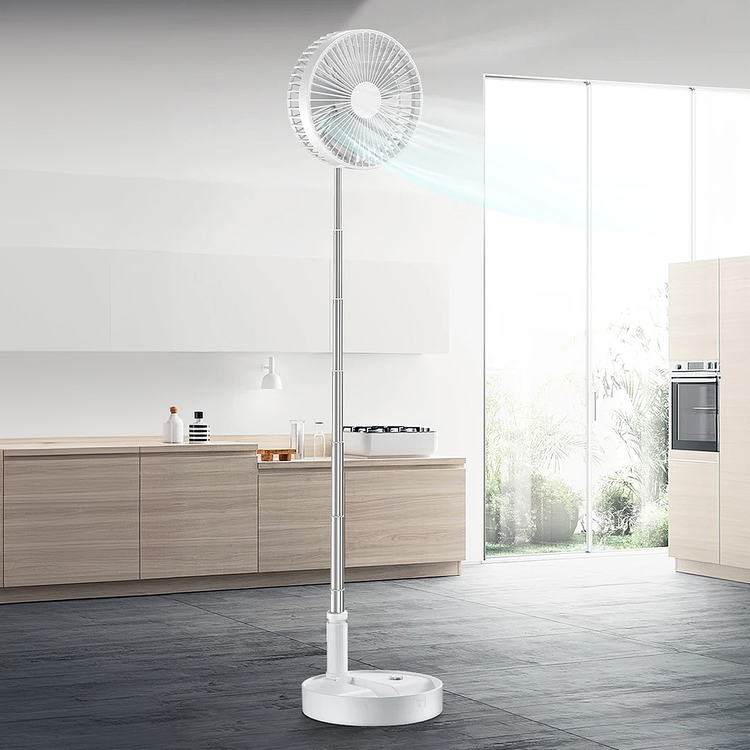 Un ventilatore bianco è acceso, che soffia aria. È in una cucina moderna con mobili in legno e una grande finestra. Il pavimento è scuro.