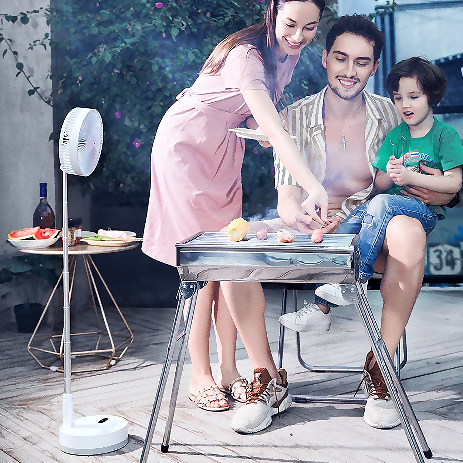 Una famiglia fa un barbecue all'aperto. La donna e l'uomo curano la griglia, un bambino è seduto in grembo all'uomo. Un ventilatore e un tavolo con cibo sono vicini.