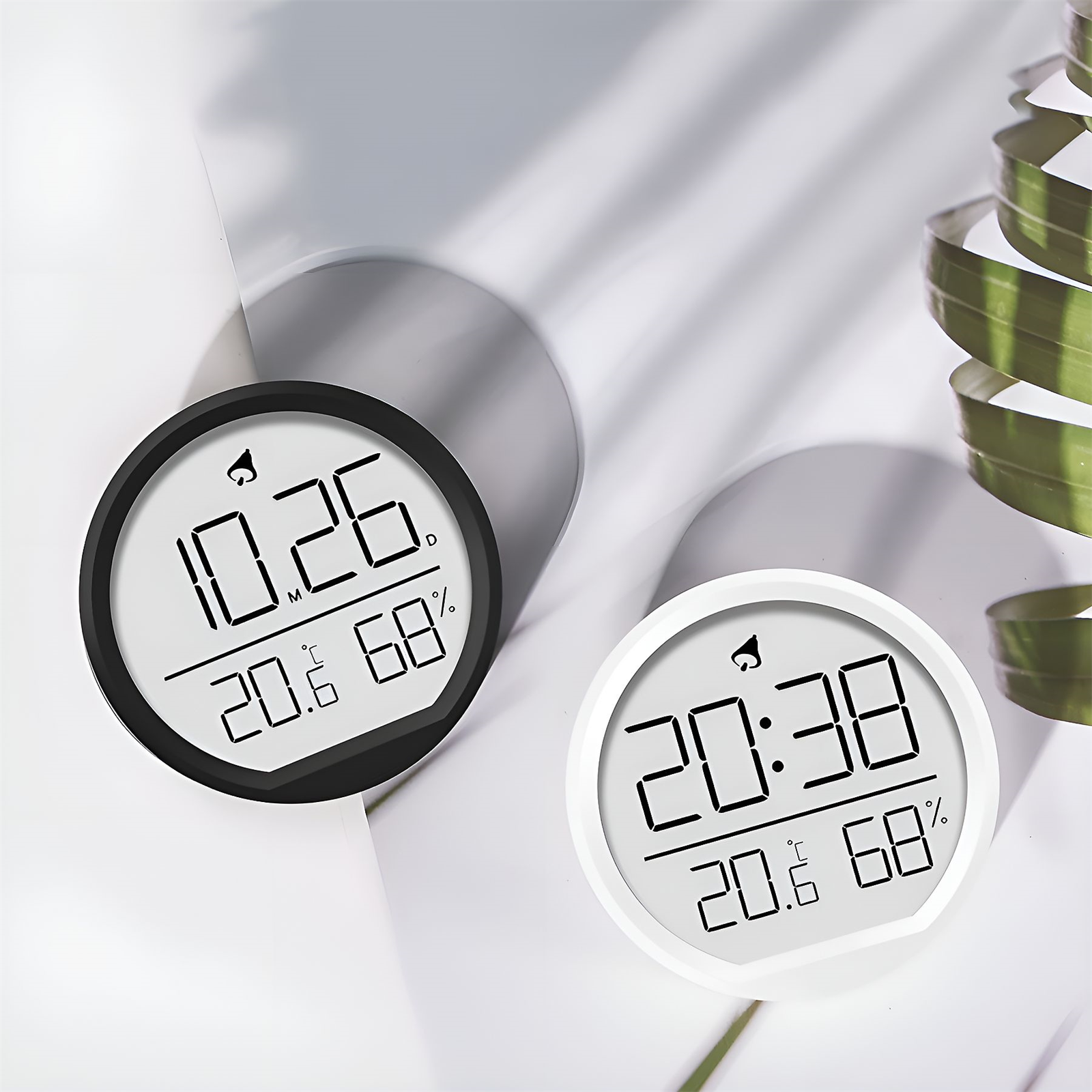 Zwei digitale Thermometer, schwarz und weiß, zeigen Zeit, Temperatur und Luftfeuchtigkeit an. Grüne Blätter im Hintergrund.
