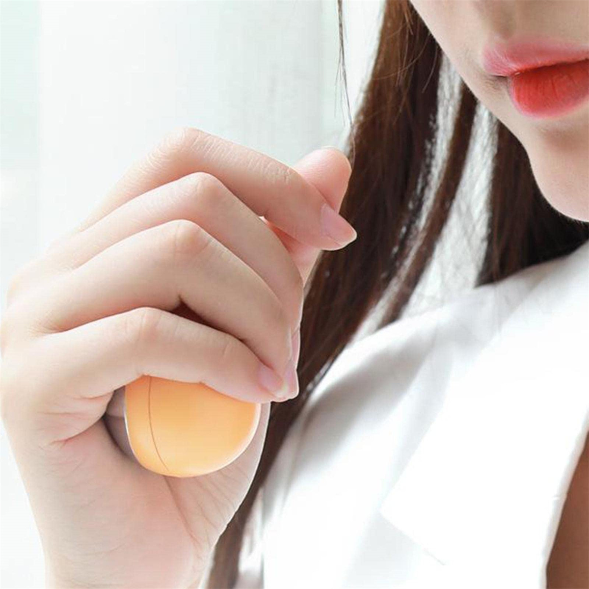 Die Hand einer Person hält einen orangefarbenen, eiförmigen Gegenstand. Die Lippen der Person sind sichtbar, rot bemalt, mit braunen Haaren.