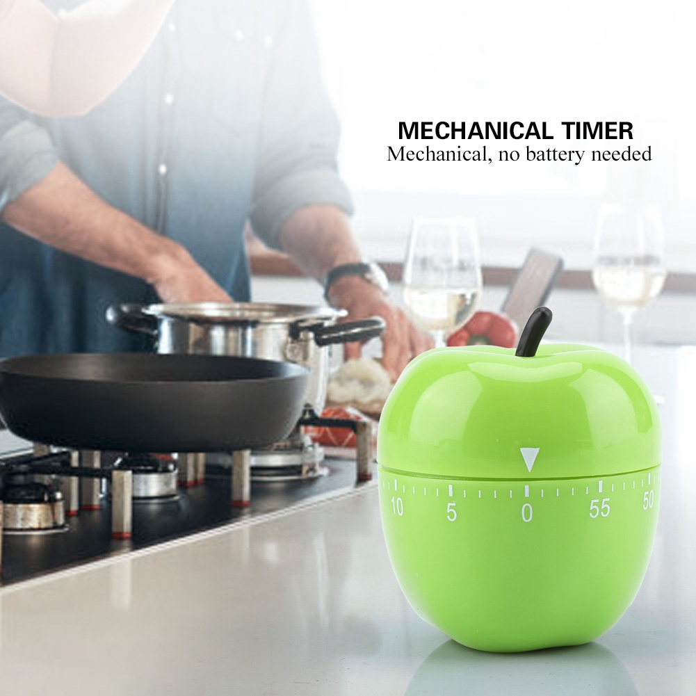 Ein grüner, apfelförmiger mechanischer Timer steht auf einer Theke. Im Hintergrund kocht eine Person. Der Timer zeigt die Zeit an.