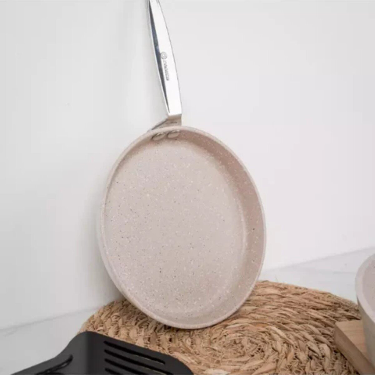 Een lichtgekleurde koekenpan met een zilveren handvat rust op een geweven placemat, naast een zwarte spatel. De achtergrond is wit.