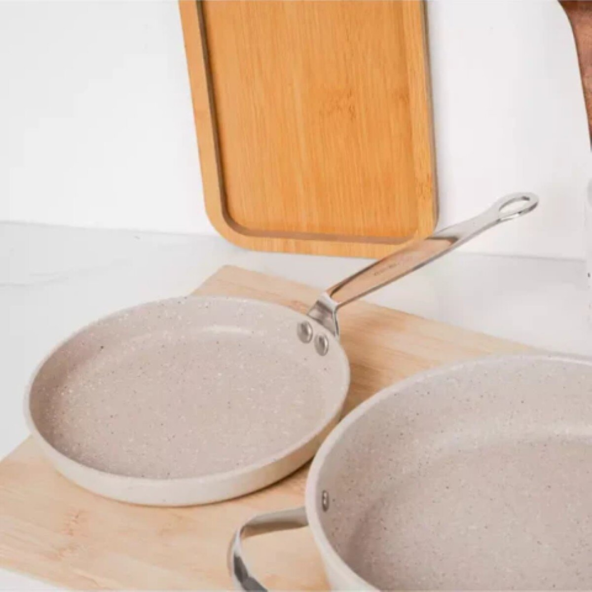 Twee lichtgrijze gespikkelde pannen met zilveren handvatten liggen op een houten snijplank. Een houten dienblad staat op de achtergrond.