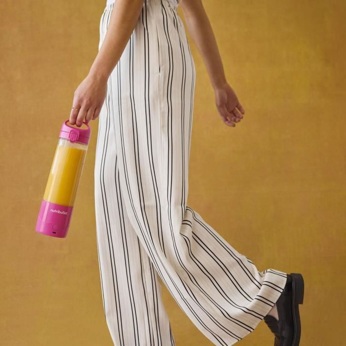 Osoba trzyma różowy Nutribullet z pomarańczowym płynem, nosi spodnie w paski i czarne buty, na żółtym tle.