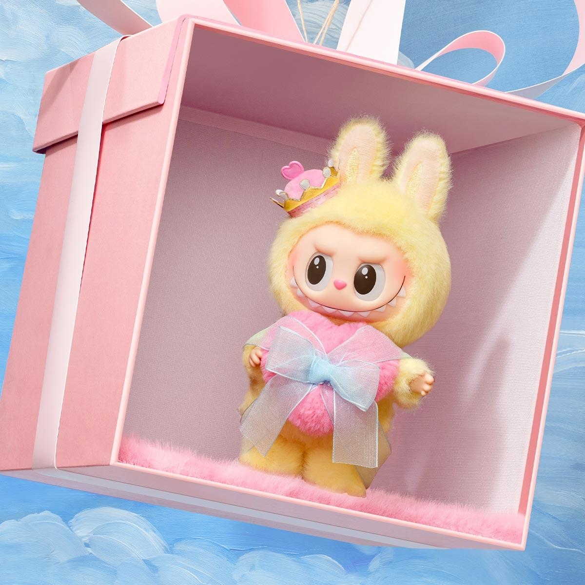 Ein Plüschtier in einer Geschenkbox. Das Spielzeug hat die Form eines Häschens und trägt eine Krone und eine Schleife. Die Box ist rosa, vor blauem Himmel.
