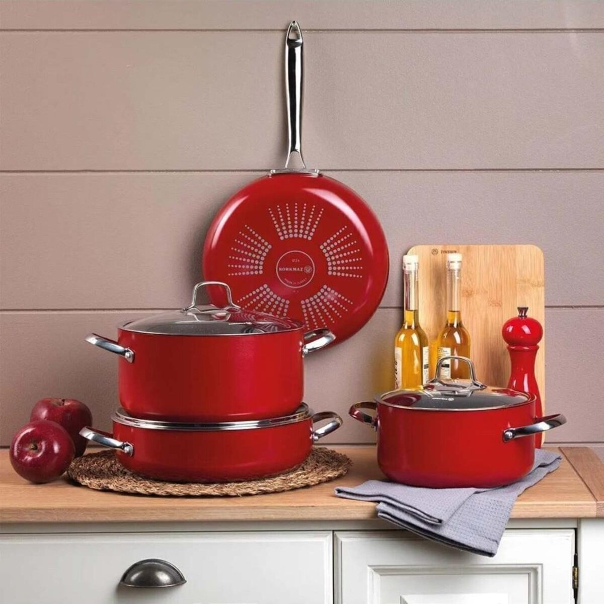 Rotes Kochgeschirr-Set mit roter Pfanne, Töpfen und Äpfeln auf einer Holzoberfläche. Hintergrund: Holzbrett, Öle und Pfeffermühle.