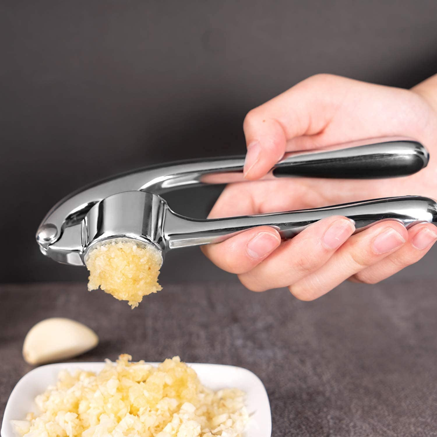 Eine Hand betätigt eine Knoblauchpresse und gibt gehackten Knoblauch auf einen Teller. Daneben liegt eine Zehe.