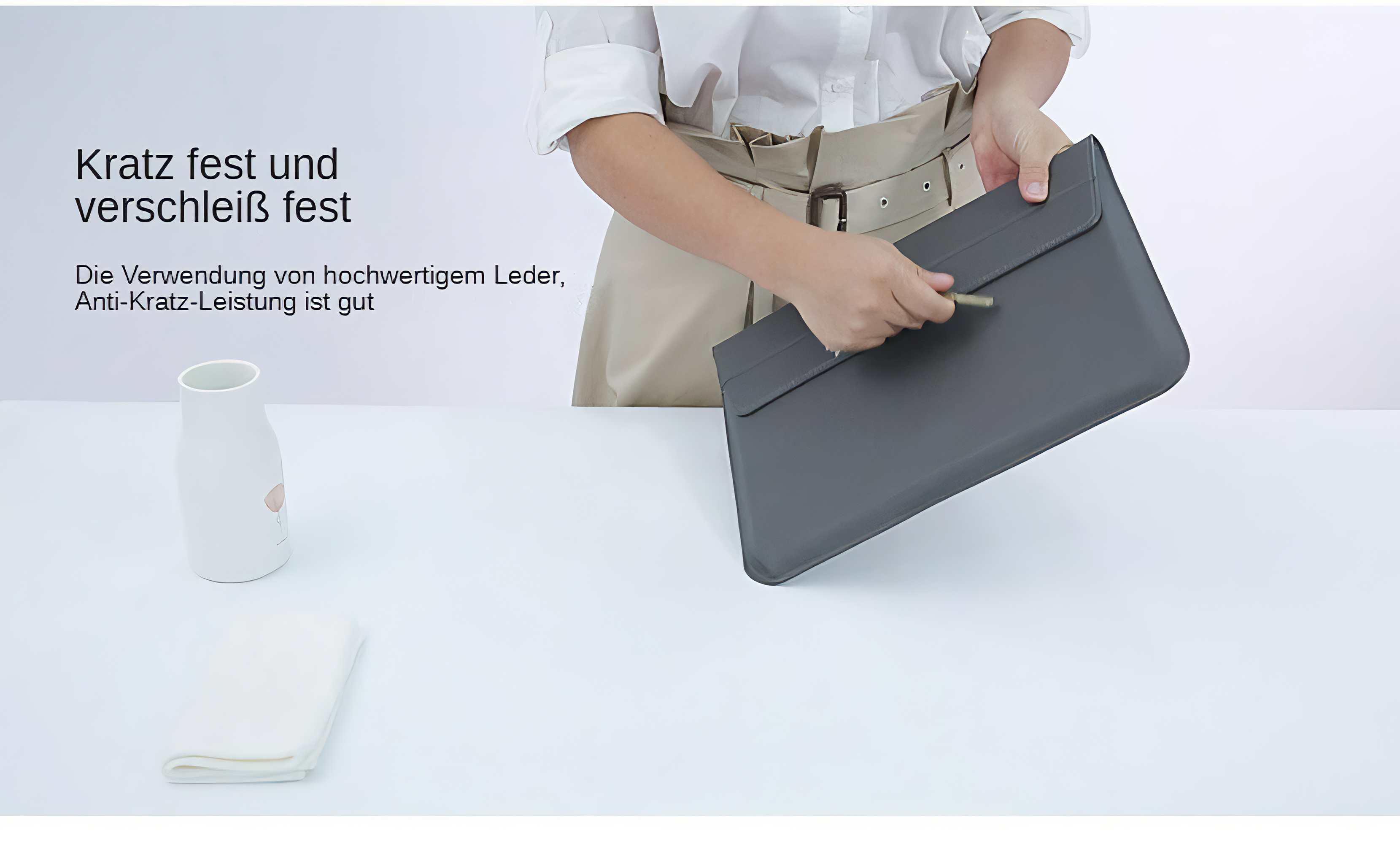 Eine Person hält eine graue Ledertasche. Daneben stehen eine weiße Vase und ein gefaltetes weißes Tuch auf einer weißen Oberfläche.