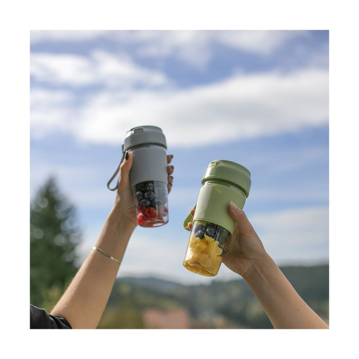 Zwei Hände halten Mixer mit Früchten vor blauem Himmel. Ein Mixer hat Blaubeeren und Himbeeren, der andere Ananas und Blaubeeren.