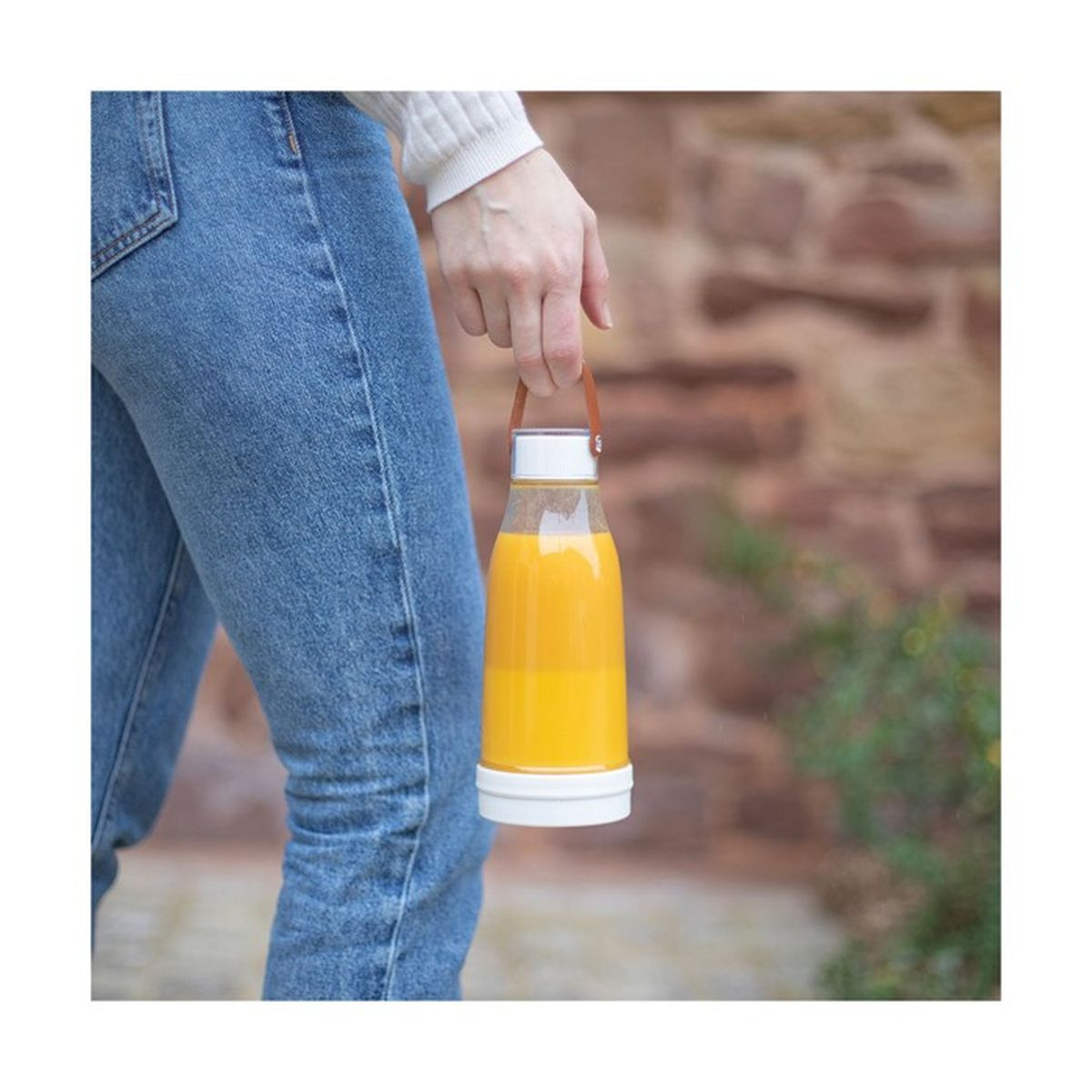Eine Person hält eine klare Flasche mit Orangensaft. Die Flasche hat einen weißen Boden und Deckel. Die Person trägt Jeans.