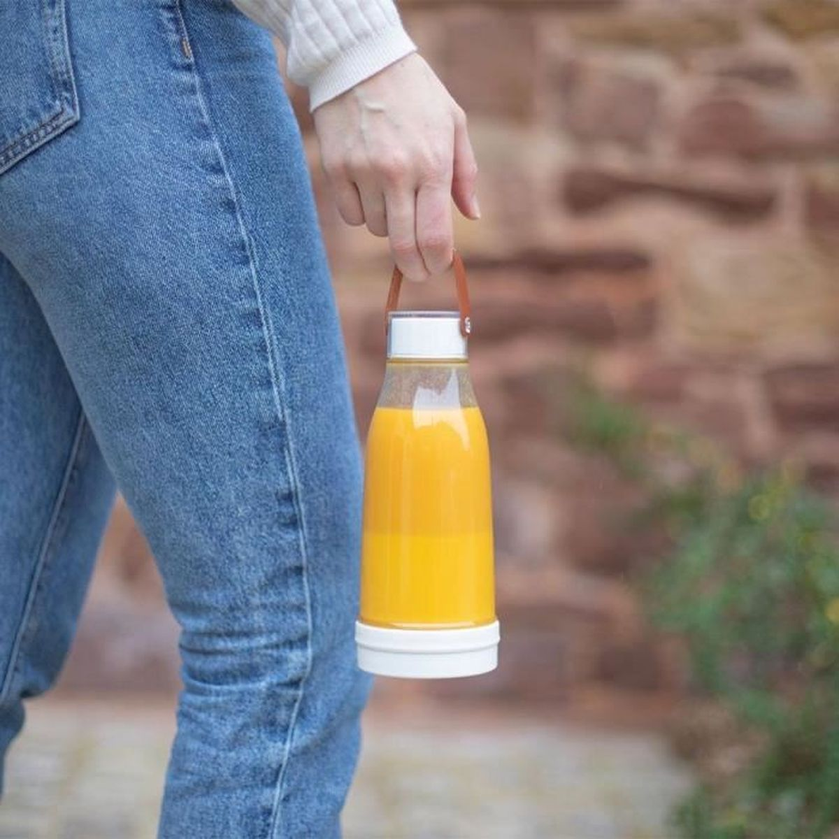 Eine Person hält eine Flasche Orangensaft. Sie trägt blaue Jeans und einen weißen Pullover. Im Hintergrund ist eine Ziegelmauer.