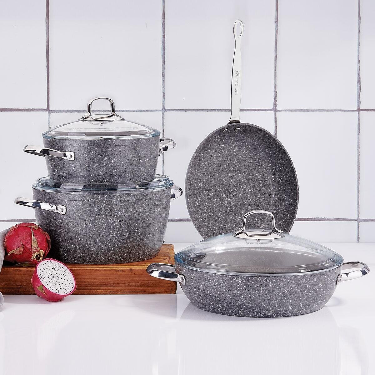 Keukengerei set. Grijs gespikkelde potten met glazen deksels en een pan met lange steel. Drakenfruit op een houten plank.