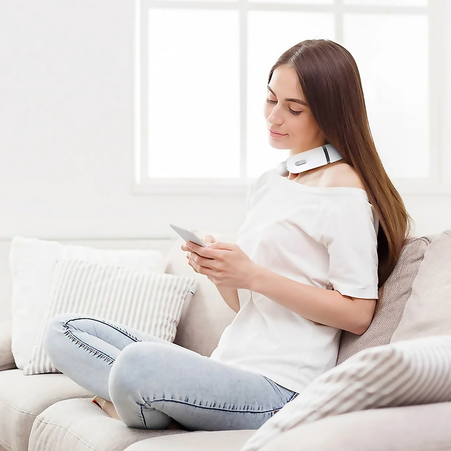 Mujer sentada en un sofá, con un masajeador de cuello, mirando un teléfono. Camisa blanca, jeans. Ventana al fondo.