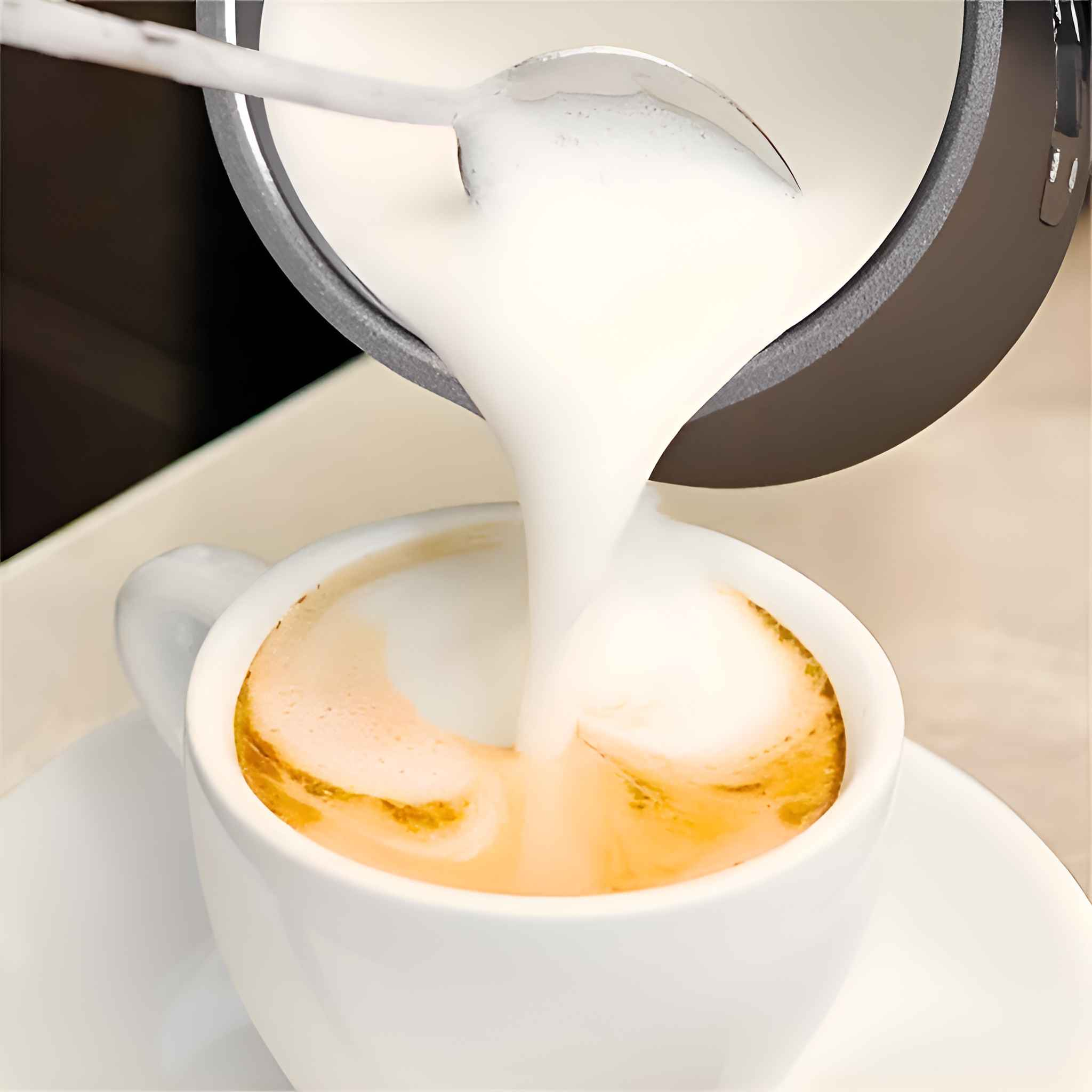 Ein Löffel gießt schaumige Milch in eine weiße Kaffeetasse mit einer hellbraunen Flüssigkeit. Die Tasse steht auf einer Untertasse.