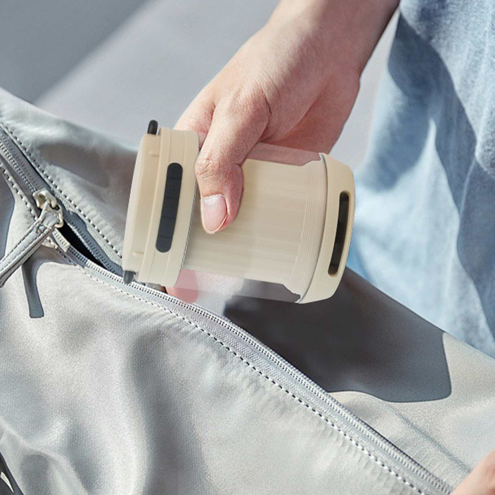 Eine Hand steckt einen beigefarbenen Behälter mit schwarzen Akzenten in eine graue Tasche. Der Reißverschluss ist teilweise geöffnet.