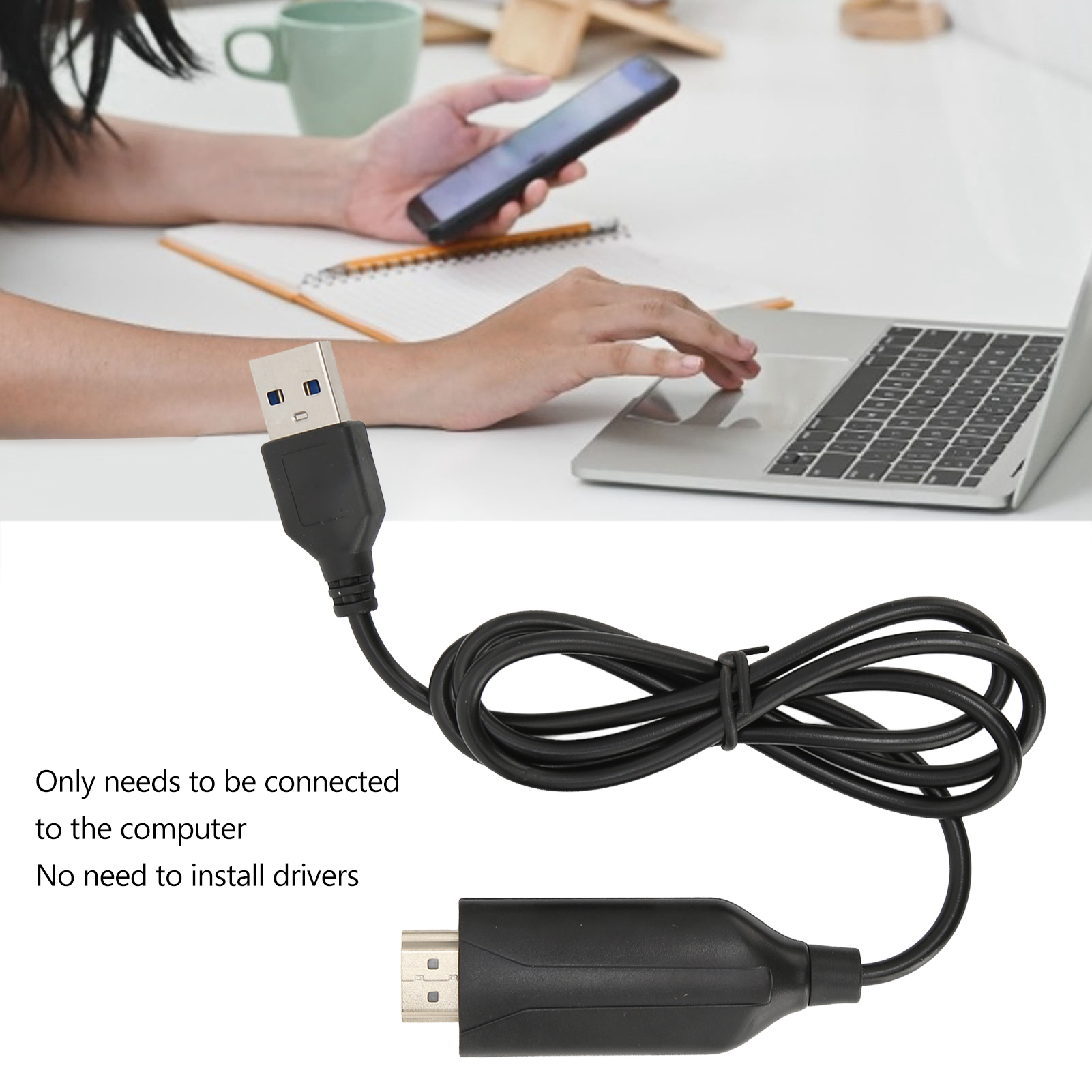 Ein USB-zu-HDMI-Kabel ist auf einer weißen Oberfläche aufgerollt. Eine Person benutzt einen Laptop und ein Telefon. Es gibt eine Tasse und ein Notizbuch.