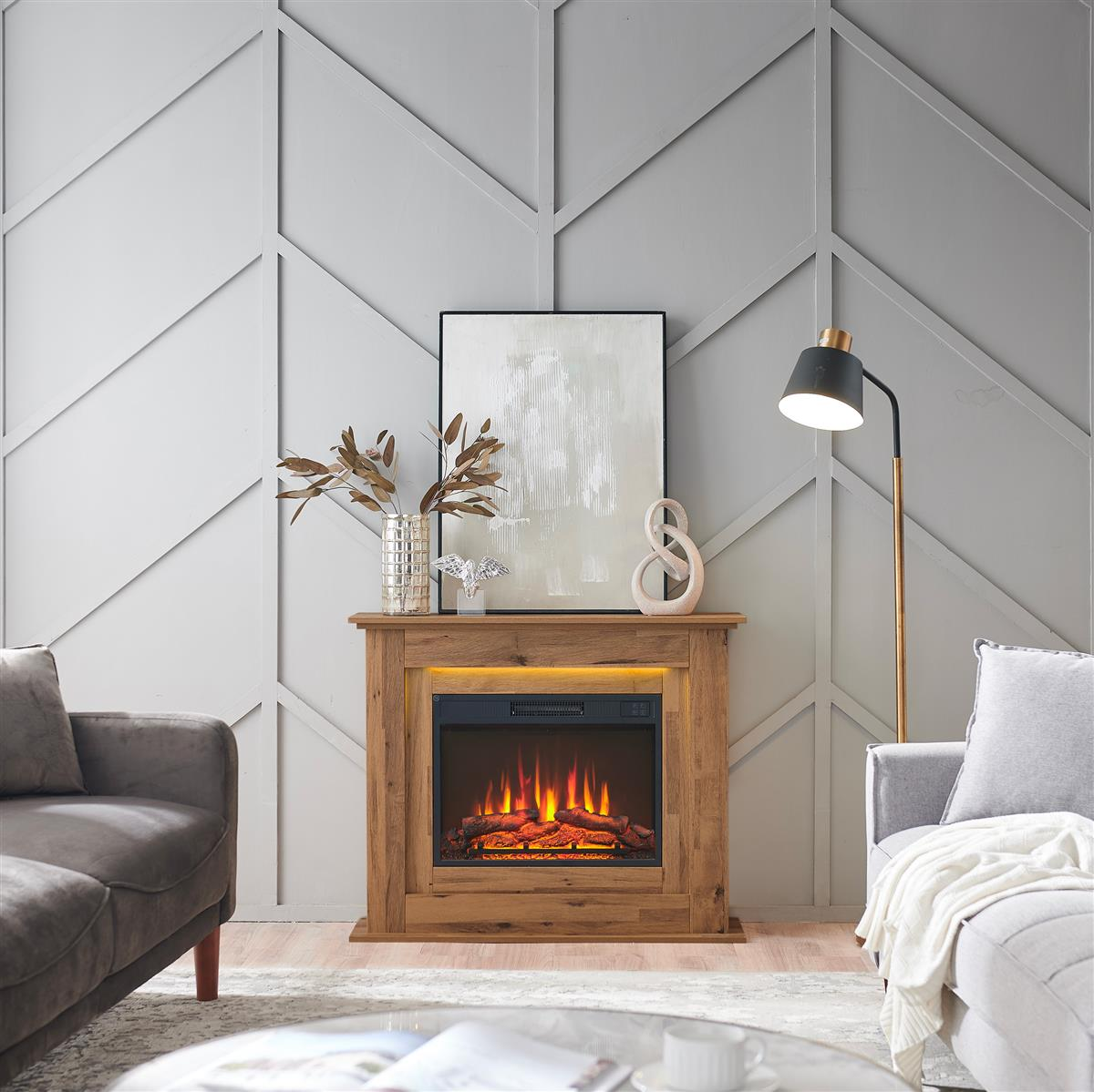 Ein Holzkamin mit Flammeneffekt steht im Mittelpunkt. Ein Gemälde, eine Vase und eine Lampe befinden sich daneben. Graue Wand und Sofas.