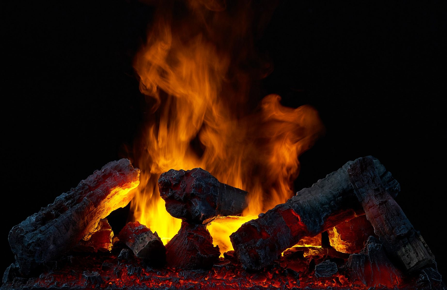 Kamin mit brennenden Holzscheiten und Flammen vor schwarzem Hintergrund. Die Holzscheite sind dunkel und die Flammen orange und gelb.