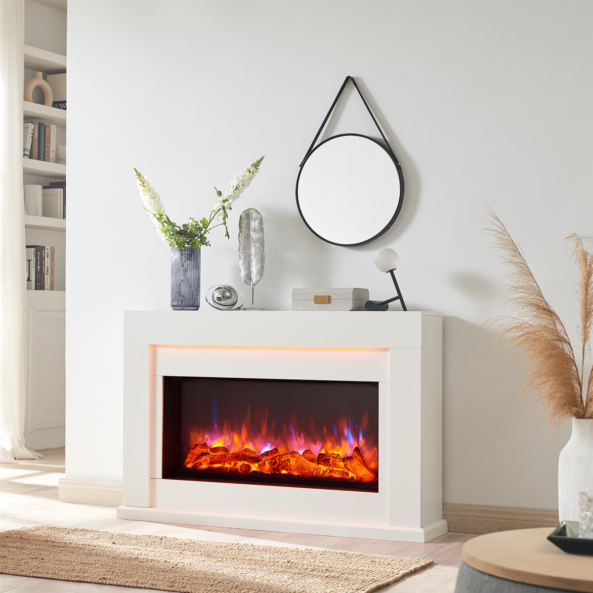 Ein weißer Kamin mit Flammendarstellung, Spiegel und Dekorationen. Auf dem Kaminsims steht eine Vase mit Blumen. Auf dem Boden liegt ein Teppich.