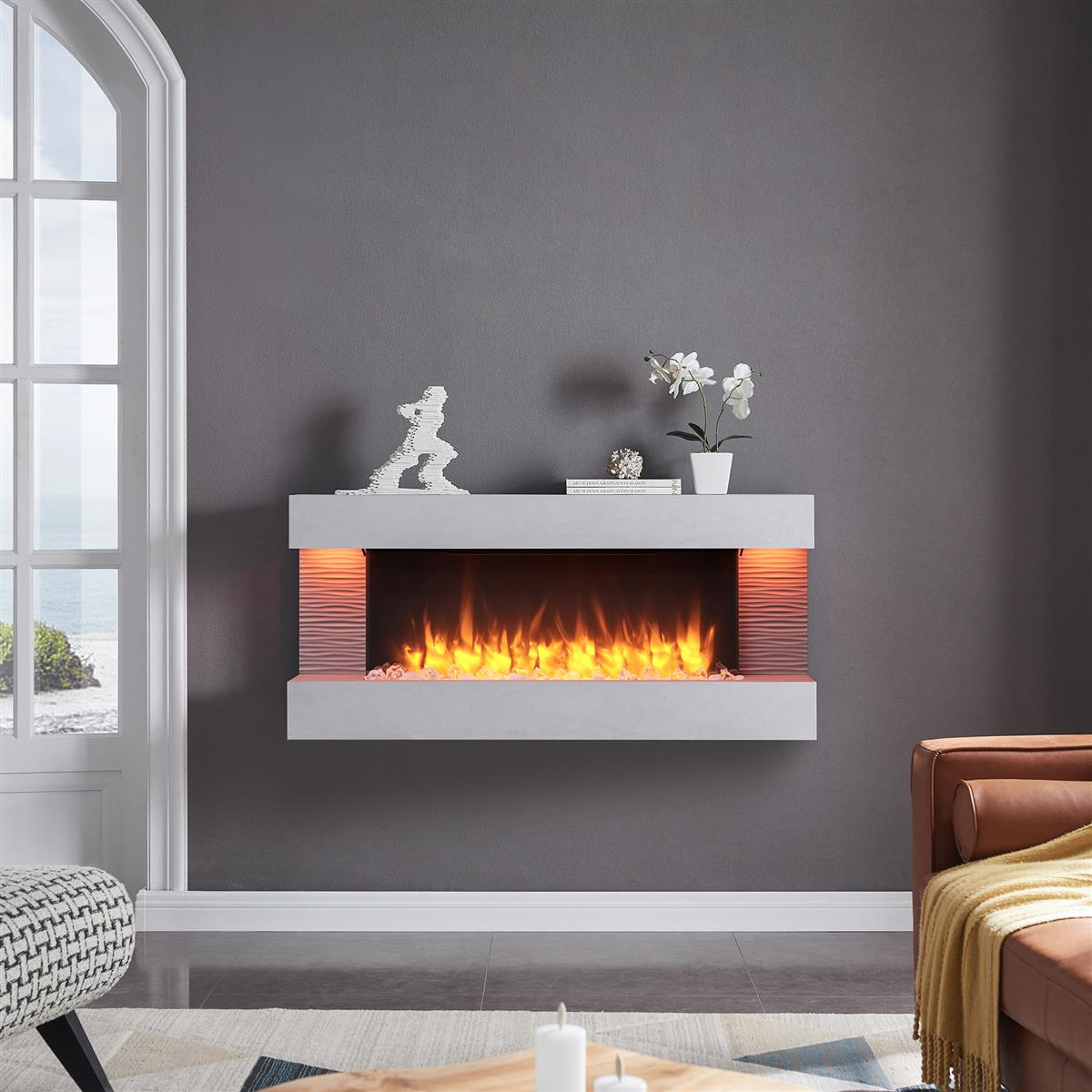 Moderner Kamin mit Flammen, weißem Mantel und Dekoration vor einer grauen Wand. Ein Fenster und eine Ledercouch sind ebenfalls sichtbar.