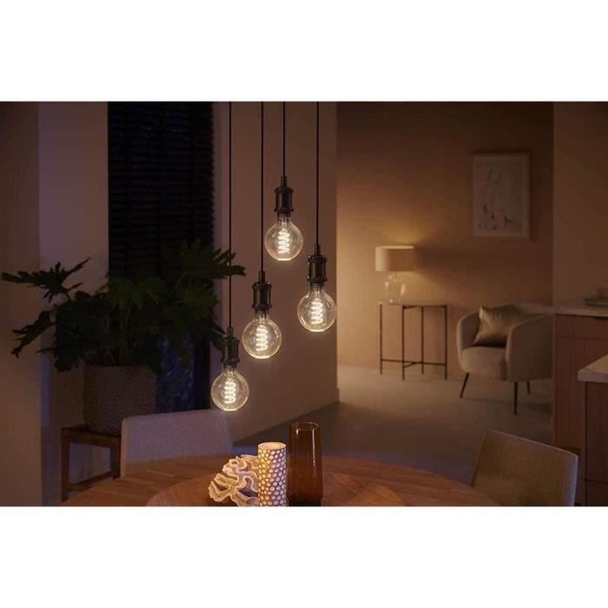 Vier hangende gloeilampen verlichten een eethoek. Een ronde houten tafel is gedekt met kaarsen. Een plant en een stoel zijn ook zichtbaar.