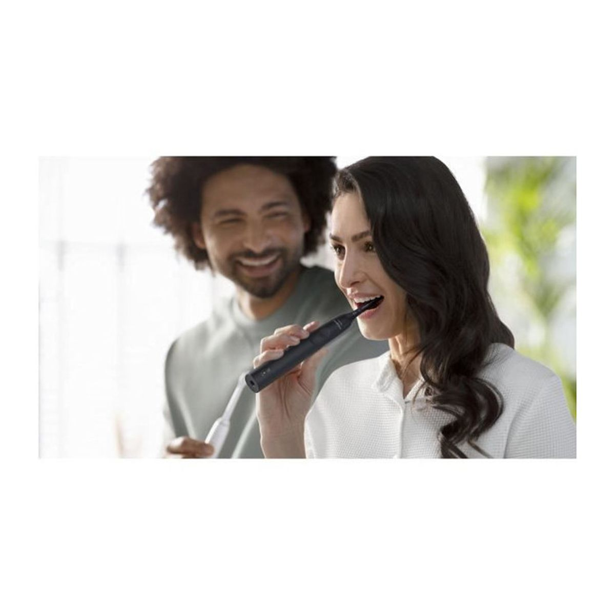 Kobieta szczotkuje zęby czarną elektryczną szczoteczką. Mężczyzna uśmiecha się w tle, trzymając szczoteczkę do zębów.