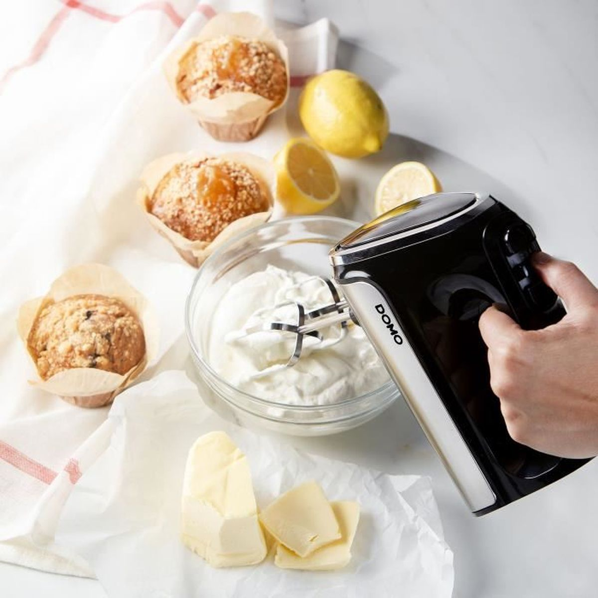 Un frullatore a mano nero mescola la panna in una ciotola di vetro. Muffin, limoni e burro sono nelle vicinanze. Una mano tiene il frullatore.