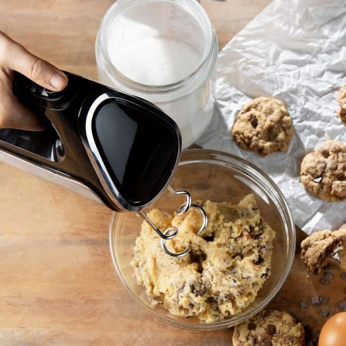 Un frullatore a mano mescola l'impasto per biscotti in una ciotola di vetro. Un barattolo di zucchero, biscotti e un uovo sono anche sulla superficie di legno.
