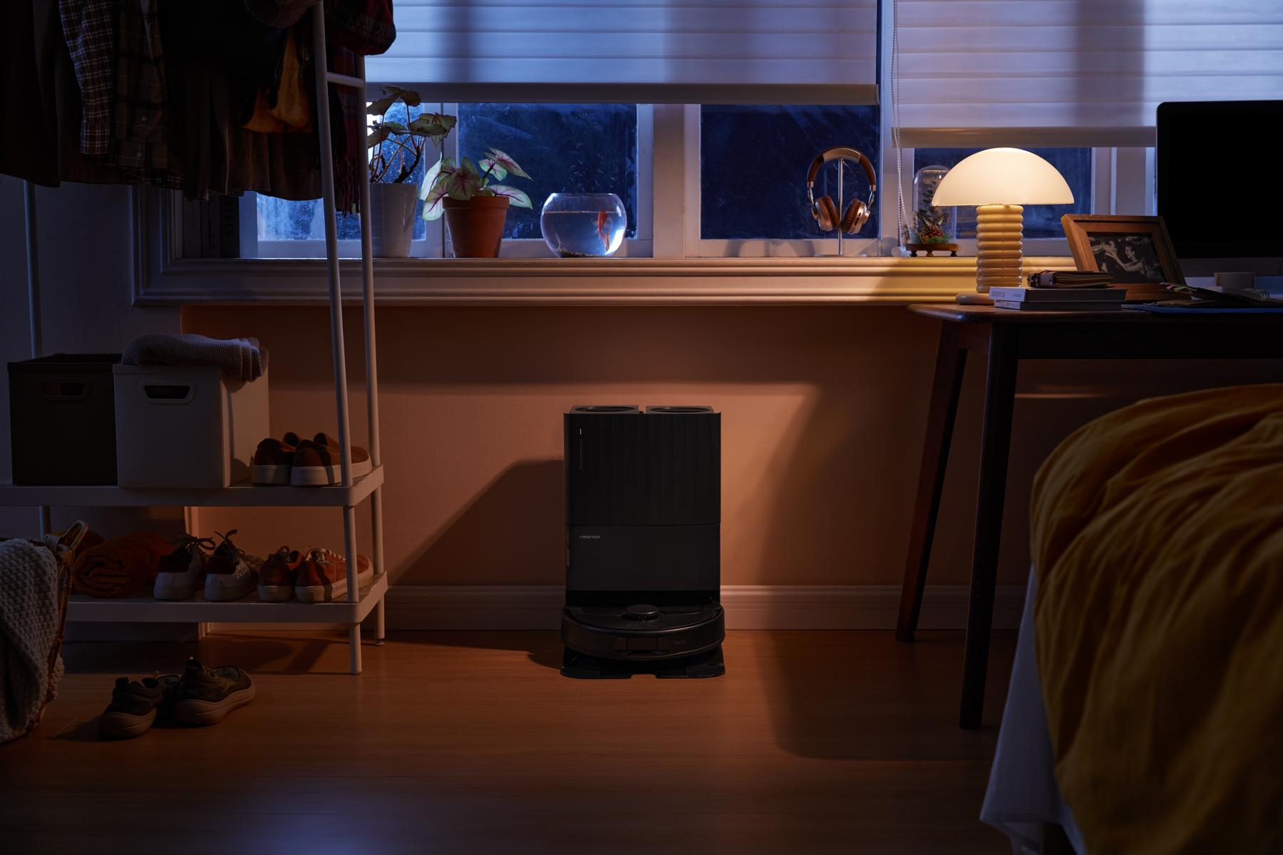 Ciemny pokój z oknem, biurkiem i czarnym robotem sprzątającym. Włączona lampka nocna. Buty i ubrania w pobliżu białego stojaka.