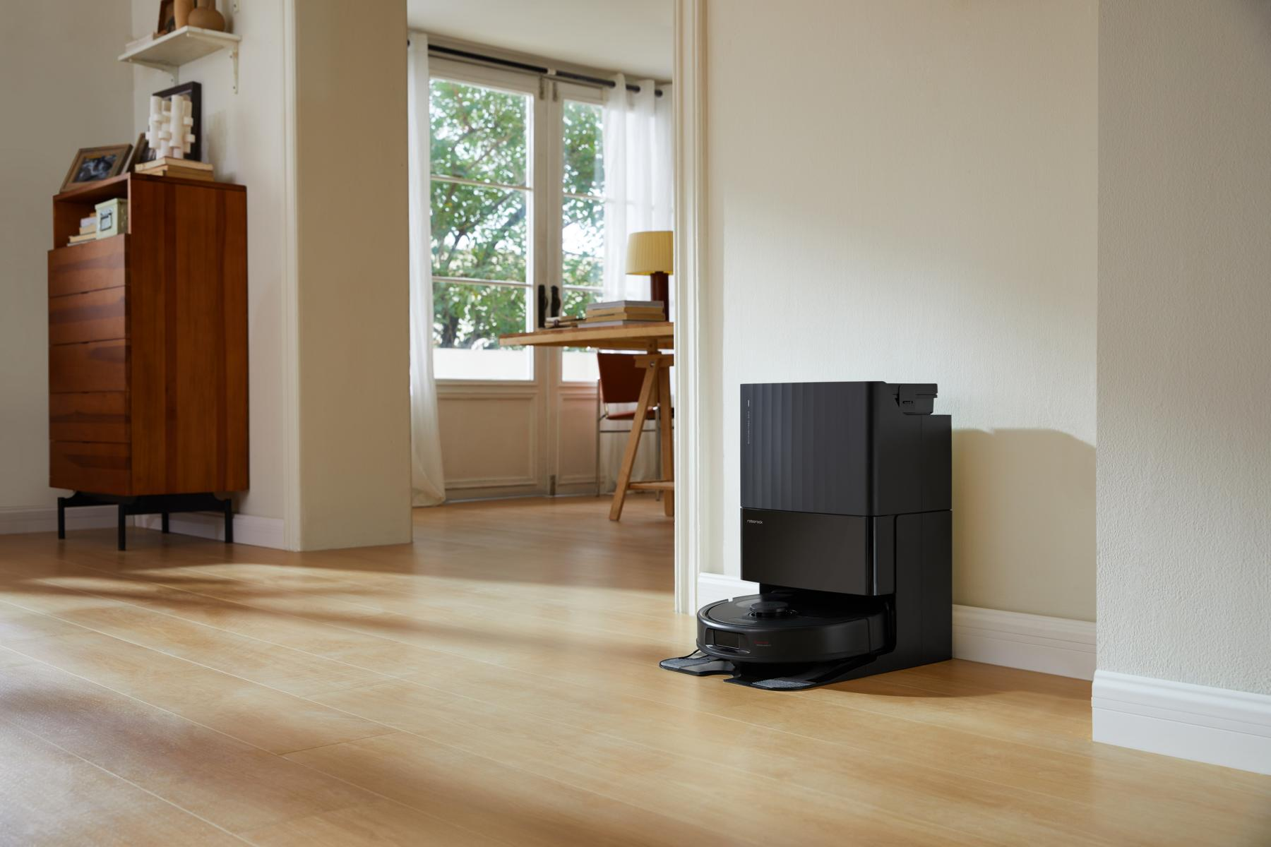 Czarny odkurzacz automatyczny stoi na drewnianej podłodze. Drewniana szafka i biurko są w tle.