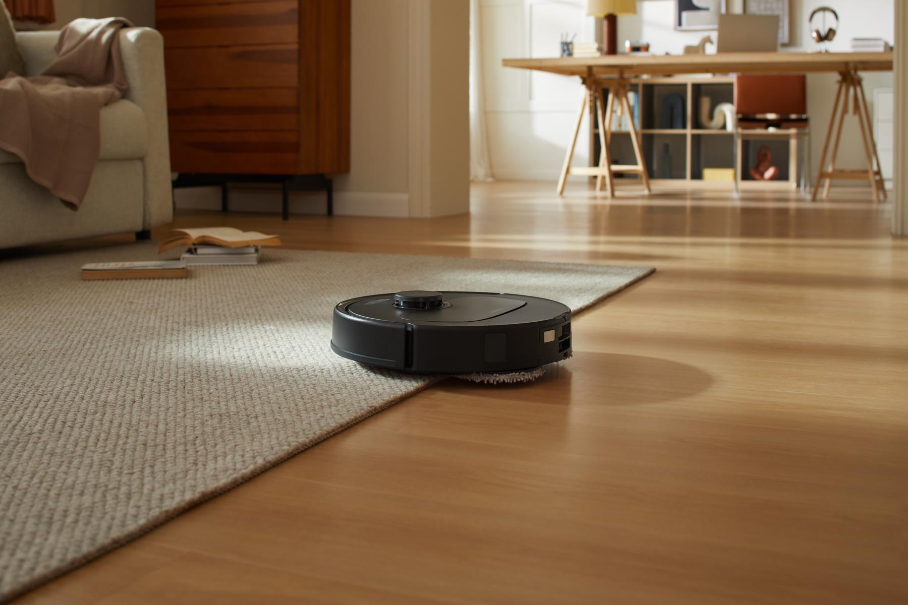 Ein schwarzer Roboter-Staubsauger auf einem Teppich. Ein Wohnzimmer mit Couch, Tisch und Holzboden im Hintergrund.