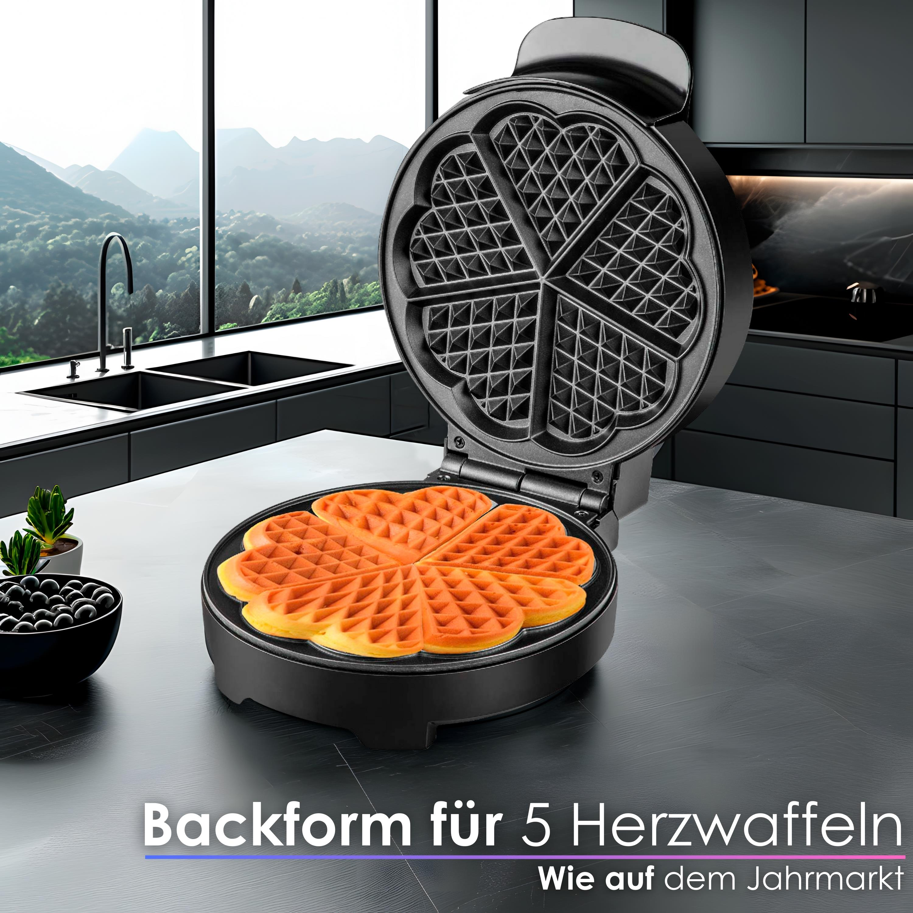Schwarzes Waffeleisen mit herzförmigen Waffeln, geöffnet. Eine Schüssel mit schwarzen Beeren und Pflanzen im Hintergrund.