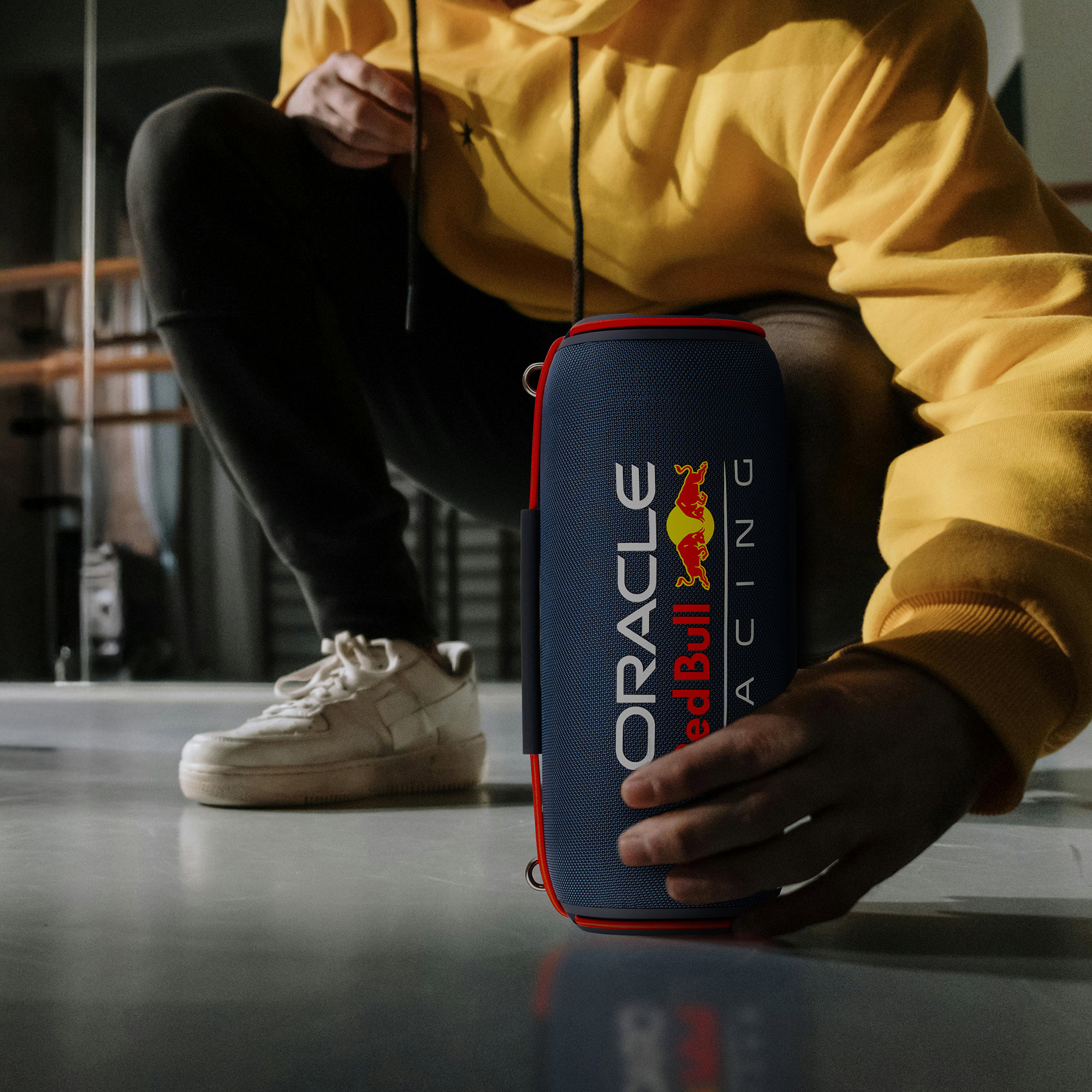 Persoon hurkt, houdt een marineblauwe luidspreker met het Oracle Red Bull Racing logo vast. Gele hoodie en witte schoenen zijn zichtbaar.
