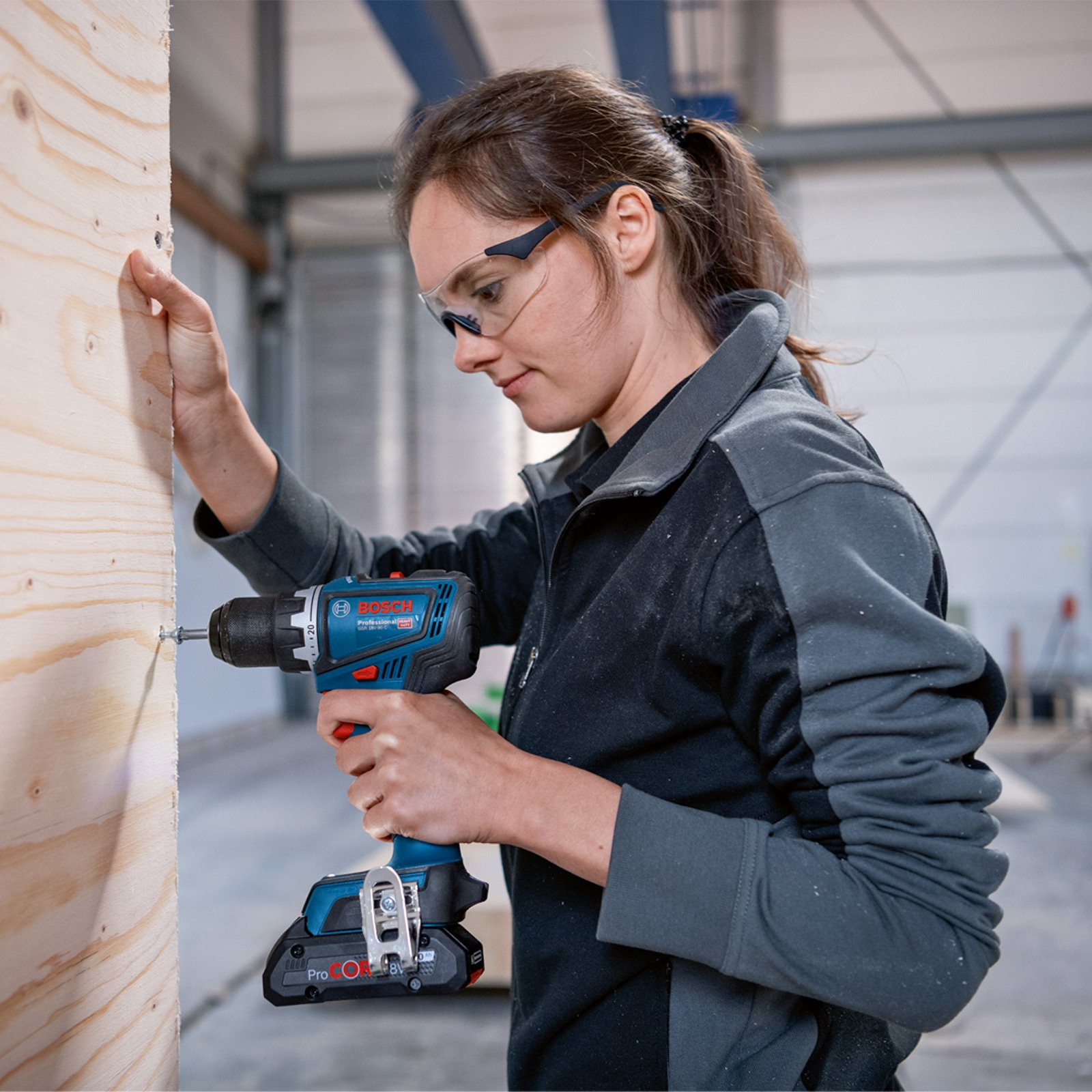 Una mujer con gafas de seguridad usa un taladro azul y negro para atornillar en una superficie de madera en un taller.