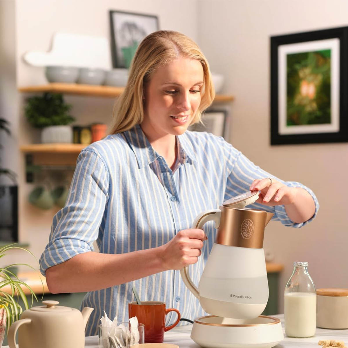 Donna bionda con un bollitore. Ambiente cucina con teiera, tazza e bottiglia di latte sul tavolo.