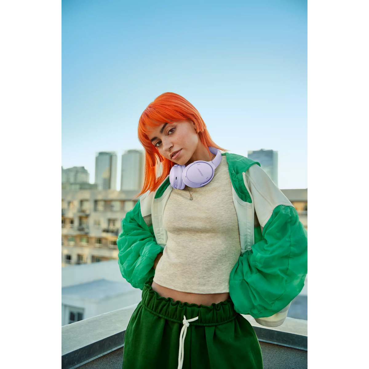 Młoda kobieta z pomarańczowymi włosami w zielonej kurtce i spodniach, słuchawki na szyi, stojąca na dachu z tłem miasta.