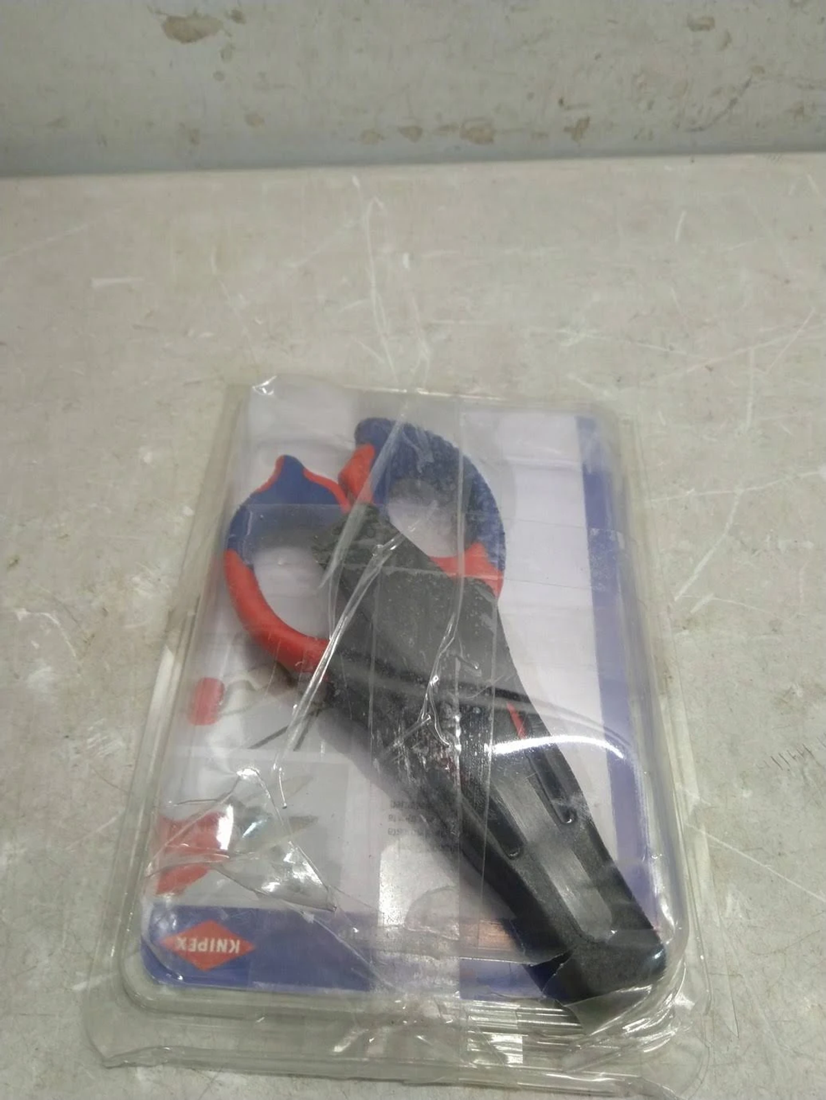Scissors in plastic packaging. Red and blue handles. Black body. The packaging is on a light gray surface.