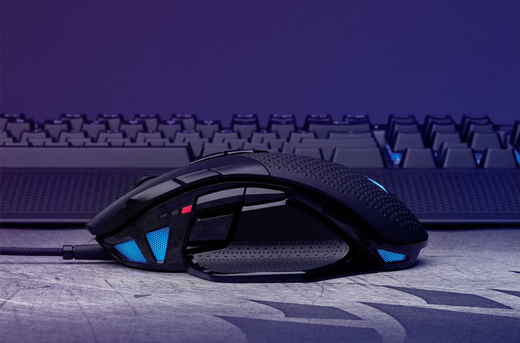 Czarna mysz do gier z niebieskimi akcentami, połączona kablem. Klawiatura w tle.