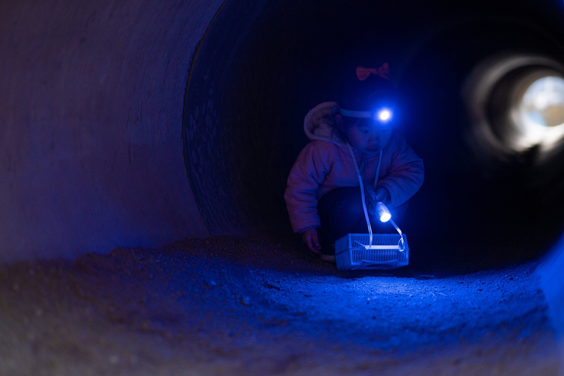 Dziecko z czołówką i latarką bada ciemny tunel. Mała klatka leży na ziemi. Niebieskie światło oświetla scenę.