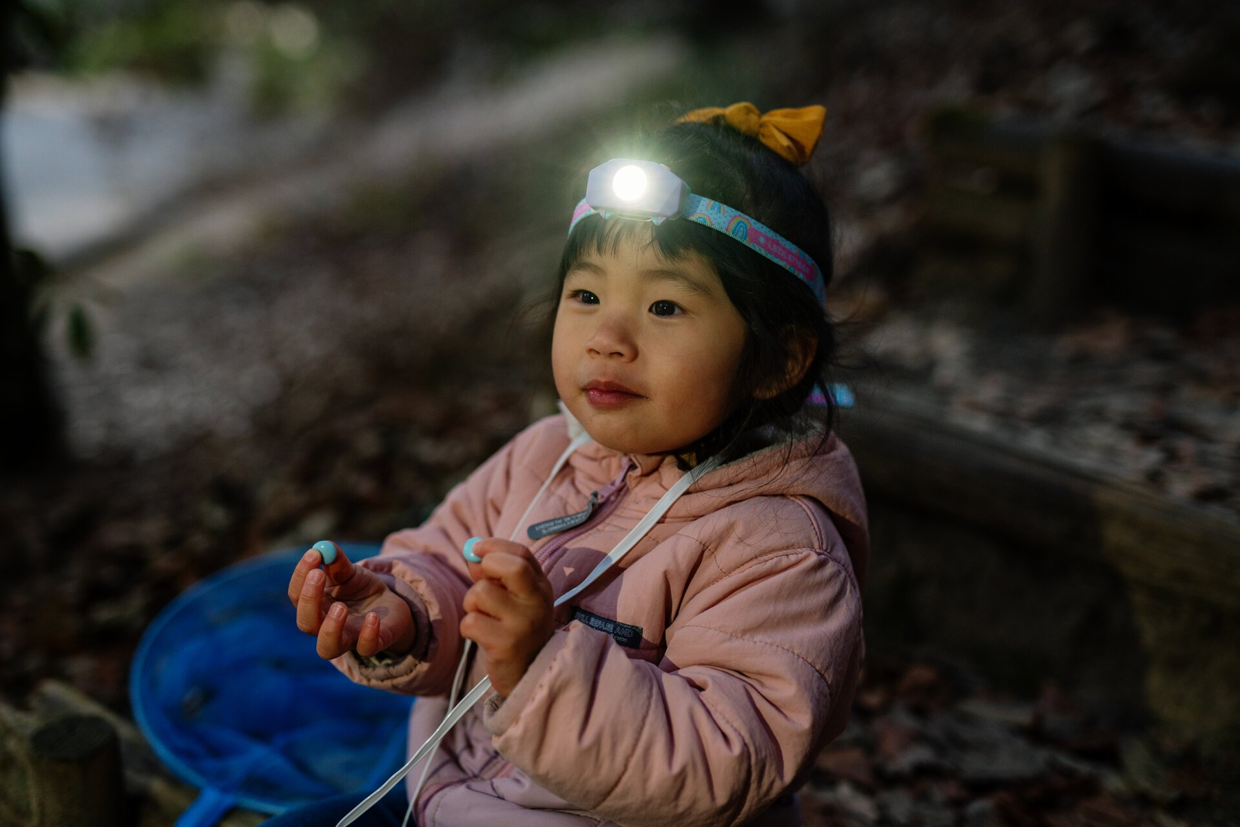 Dziecko w lampie czołowej trzyma niebieskie przedmioty. Ma na sobie różową kurtkę i kokardkę. Tło to rozmyty las.