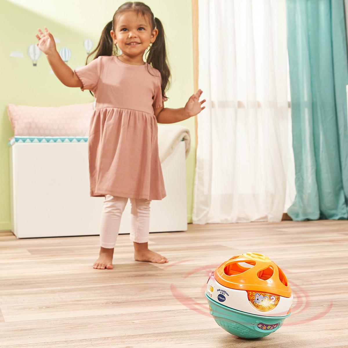 Ein junges Mädchen in rosa Kleid und Leggings lächelt, die Arme ausgestreckt. Ein bunter Ball liegt auf dem Boden. Heller Holzboden und weißer Hintergrund.