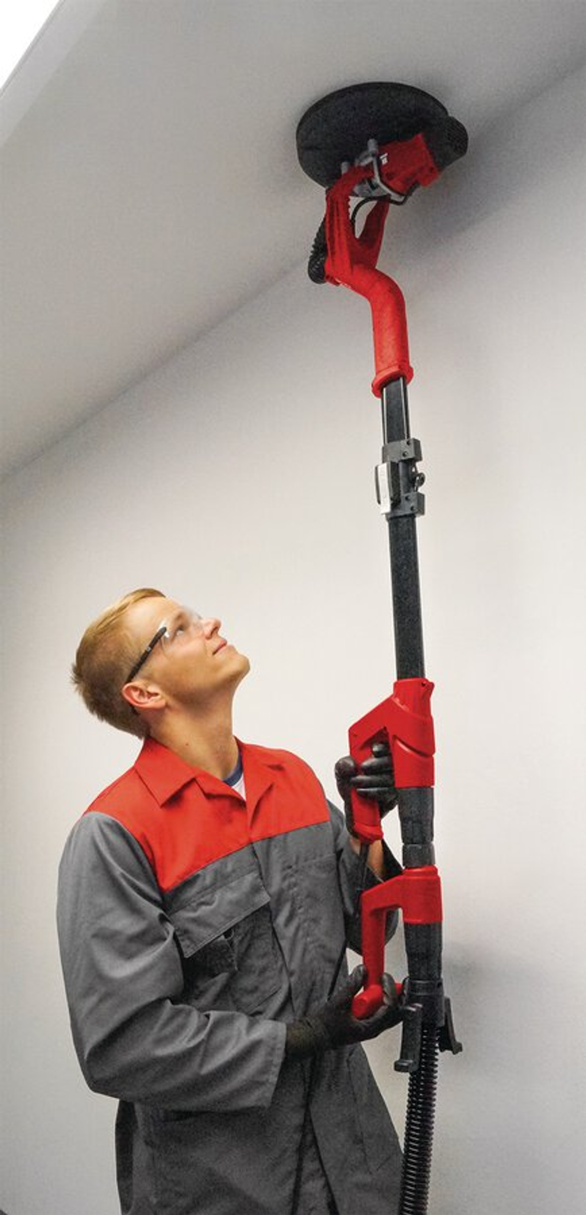 Un homme en tenue de travail ponce un plafond avec une ponceuse à long manche. Il porte des lunettes de sécurité et regarde en l'air. La ponceuse est rouge et noire.