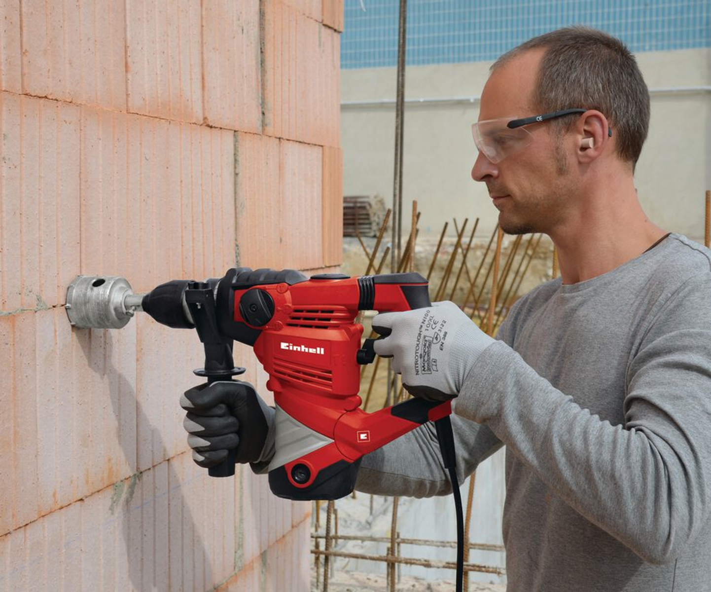 Un uomo fora un muro di mattoni con un trapano Einhell rosso e nero. Indossa occhiali e guanti di sicurezza.