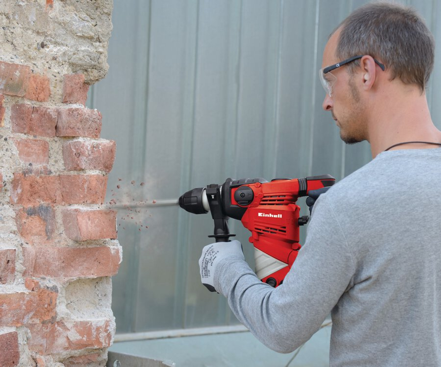 Un uomo fora un muro di mattoni con un trapano rosso Einhell. Indossa occhiali e guanti di sicurezza. Lo sfondo è un pannello metallico.