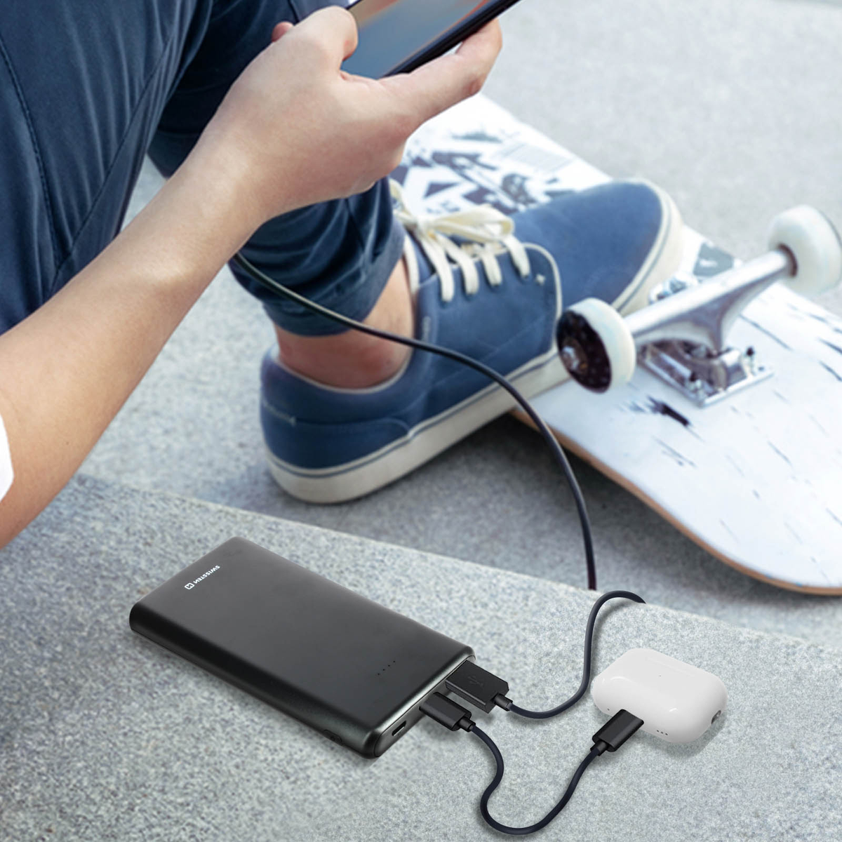 Person benutzt Handy, lädt von Powerbank und Ohrhörern. Skateboard und blaue Schuhe im Hintergrund. Schwarze Kabel verbinden Geräte.