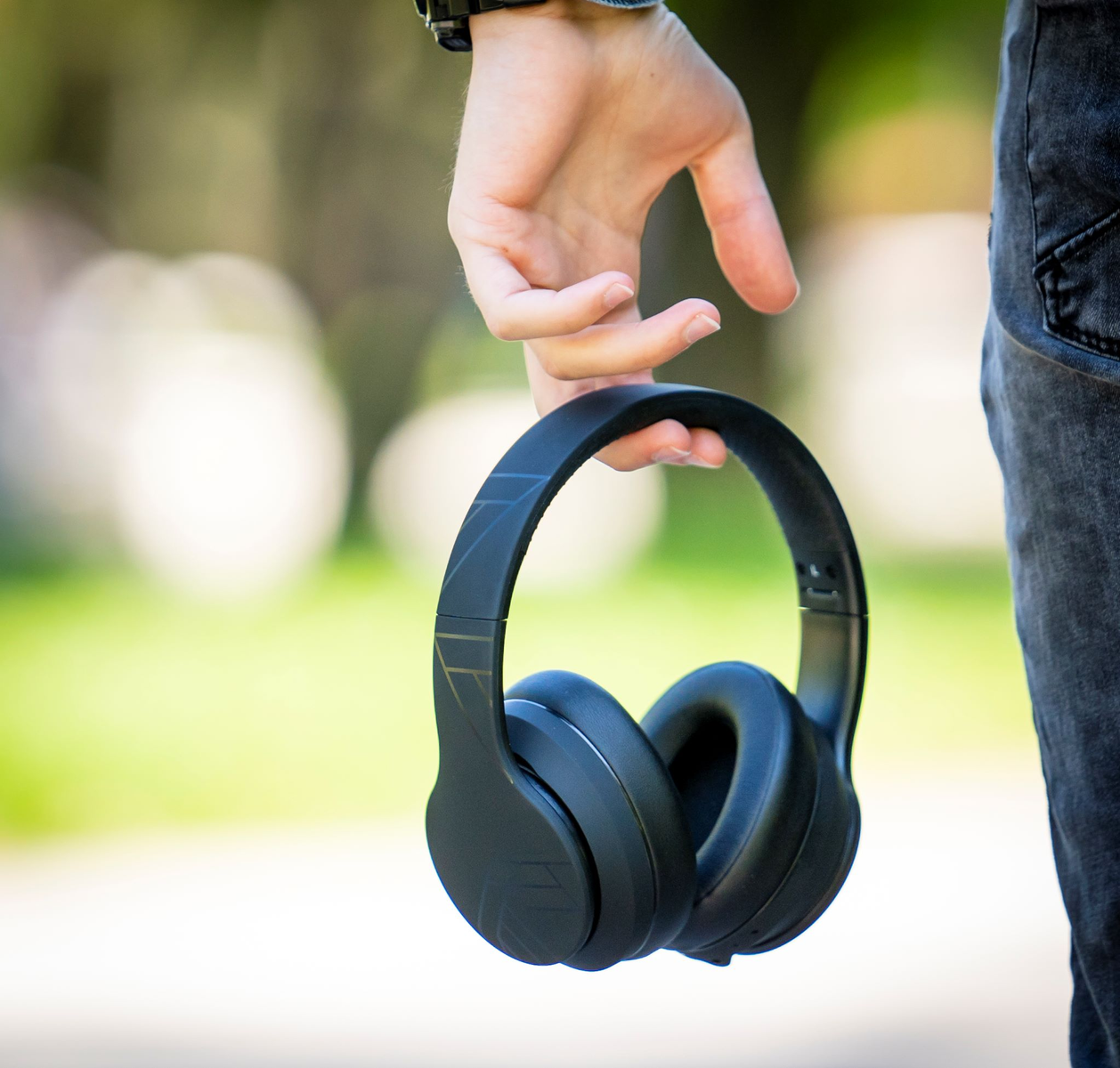 Una persona sostiene auriculares negros. El fondo es follaje verde borroso. La persona lleva un reloj y vaqueros.